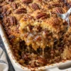 A spoon lifts a scoop of pecan pie dump cake from a white 9x13 dish, gooey pecan filling dripping under a golden cake mix crust with toasted pecans.