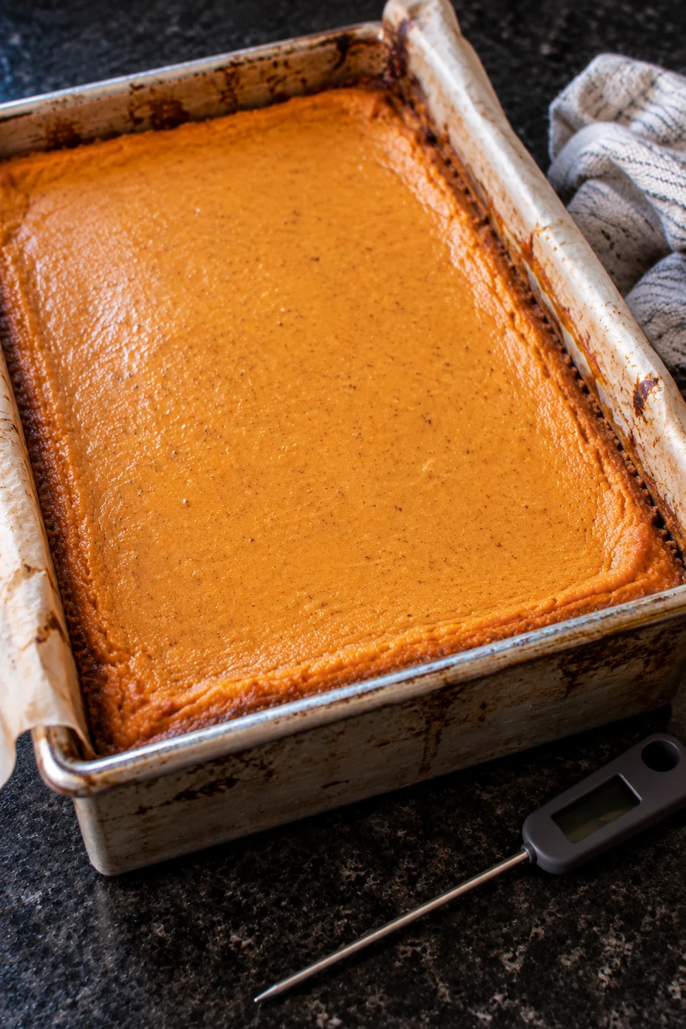 A 9x13 pan of baked pumpkin cheesecake with a smooth, crack-free top and set edges, parchment overhang on the sides and an instant-read thermometer beside it.