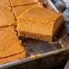 A metal spatula lifting a pumpkin cheesecake bar out of a 9x13 pan on a black stone counter, showing a graham cracker crust under a thick, smooth pumpkin cheesecake layer.