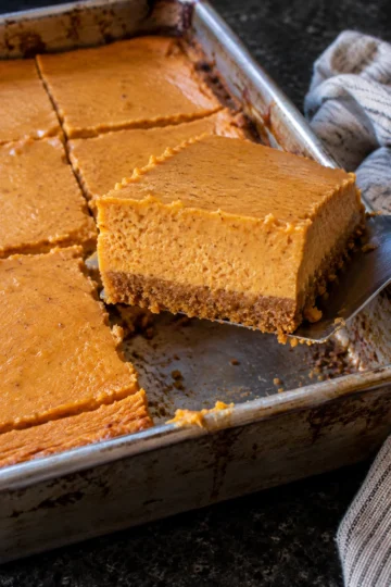 A metal spatula lifting a pumpkin cheesecake bar out of a 9x13 pan on a black stone counter, showing a graham cracker crust under a thick, smooth pumpkin cheesecake layer.