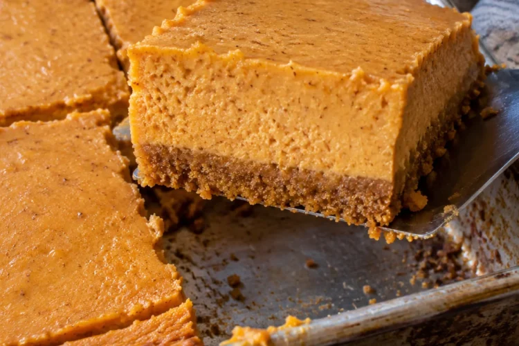 A metal spatula lifting a pumpkin cheesecake bar out of a 9x13 pan on a black stone counter, showing a graham cracker crust under a thick, smooth pumpkin cheesecake layer.