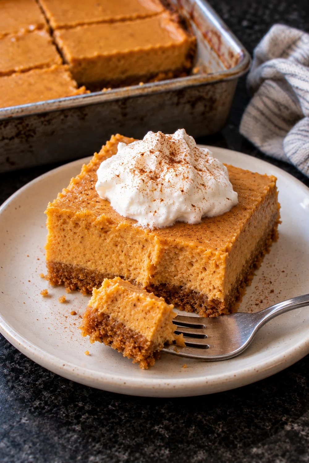 A pumpkin cheesecake bar on a cream plate topped with whipped cream and cinnamon, a fork holding one bite with crust, and the pan of bars behind.