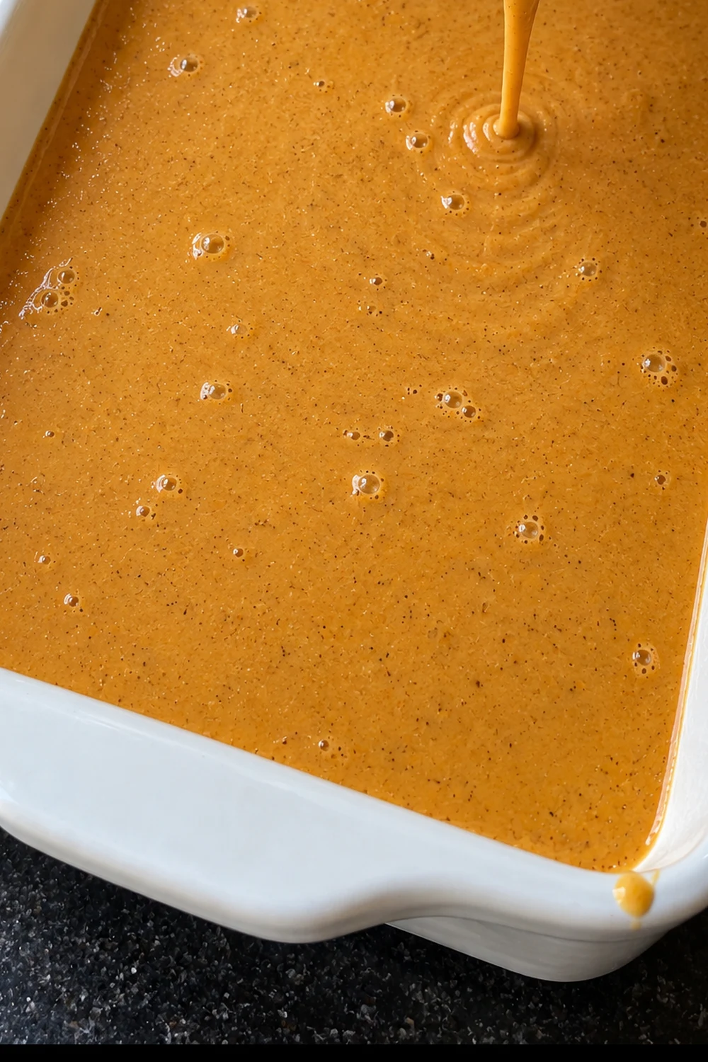 Orange pumpkin custard being poured into a greased white 9x13 baking dish in a level layer, a ribbon of custard falling into the center.