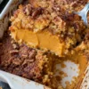A spoon lifts a scoop of pumpkin dump cake from a white 9x13 dish, showing a thick, creamy pumpkin pie layer under a golden cake mix crust with pecans.
