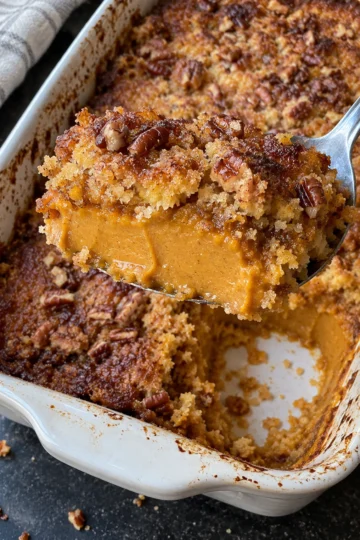 A spoon lifts a scoop of pumpkin dump cake from a white 9x13 dish, showing a thick, creamy pumpkin pie layer under a golden cake mix crust with pecans.