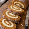 A pumpkin roll with cream cheese filling on a wooden board on a black stone counter, cut into thick slices that show the orange and white spiral, with a serrated knife beside it.