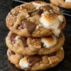 A stack of three s'mores cookies on a black stone counter, topped with toasted marshmallows, melted milk chocolate and graham cracker pieces, with more cookies cooling on a wire rack behind.