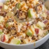A spoonful of Snickers apple salad lifted from a white serving bowl, with green and red apple chunks, Snickers pieces showing peanuts and caramel, creamy vanilla dressing and a caramel drizzle, on a black stone counter.