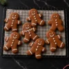 Seven soft gingerbread men on a wire cooling rack over parchment, each piped by hand with white royal icing cuffs, eyes and buttons, a pine sprig and red and white twine on the black marble around them.