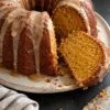 A glazed sweet potato pound cake on a speckled cream plate with one thick slice cut and tipped on its side, showing a moist golden-orange crumb under brown butter glaze drips, on a black stone counter.