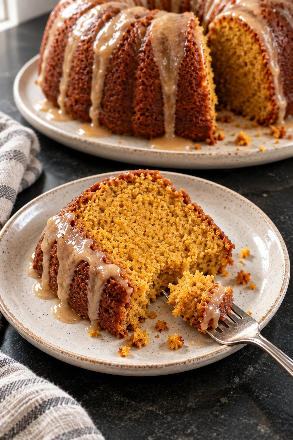 A slice of glazed sweet potato pound cake on a speckled plate with a fork holding one bite of the soft orange crumb, and the rest of the bundt cake behind.