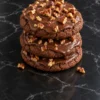 A stack of three soft chocolate Texas sheet cake cookies on black marble, each covered in glossy fudge icing that drips over the edge, topped with chopped toasted pecans, with two pecan pieces on the counter.