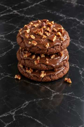 A stack of three soft chocolate Texas sheet cake cookies on black marble, each covered in glossy fudge icing that drips over the edge, topped with chopped toasted pecans, with two pecan pieces on the counter.